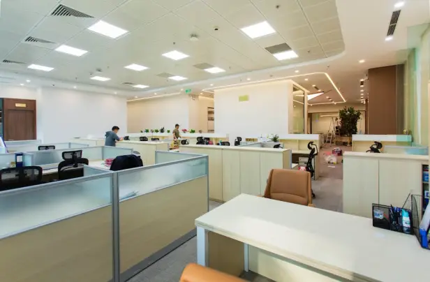 Interior de oficina luminoso y espacioso con cubículos diáfanos, mobiliario moderno e iluminación empotrada en el techo.