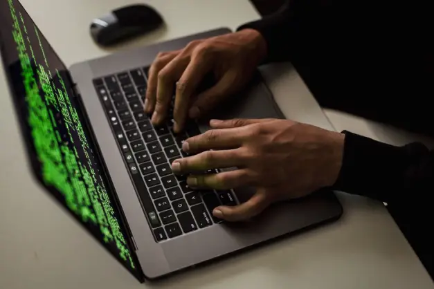 Primer plano de manos tecleando código en un portátil con texto verde en la pantalla, sugiriendo hackeo o programación. (Foto: Pexels)