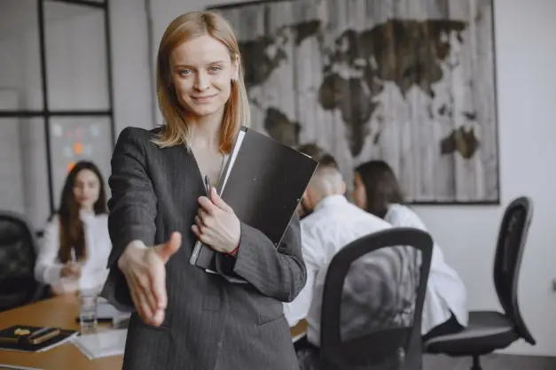 Mujer profesional ofreciendo un apretón de manos en un entorno de oficina vibrante, simbolizando trabajo en equipo y colaboración. (Foto: Pexels)