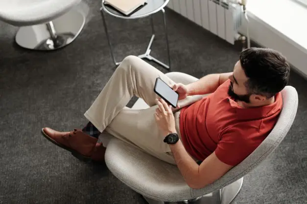 Hombre con barba en una camisa polo roja sentado cómodamente, usando un smartphone en interiores. (Foto: Pexels)