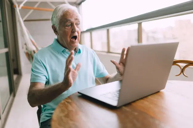 Hombre mayor luciendo sorprendido mientras usa un laptop en un balcón, mostrando emoción o sorpresa. (Foto: Pexels)