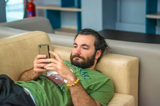 Hombre con barba tumbado cómodamente en un sofá, absorto en su smartphone en el interior. (Foto: Pexels)
