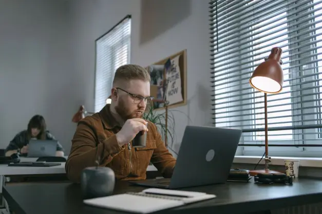 Un hombre con barba bebiendo café mientras trabaja en un portátil en un espacio de oficina moderno. (Foto: Pexels)