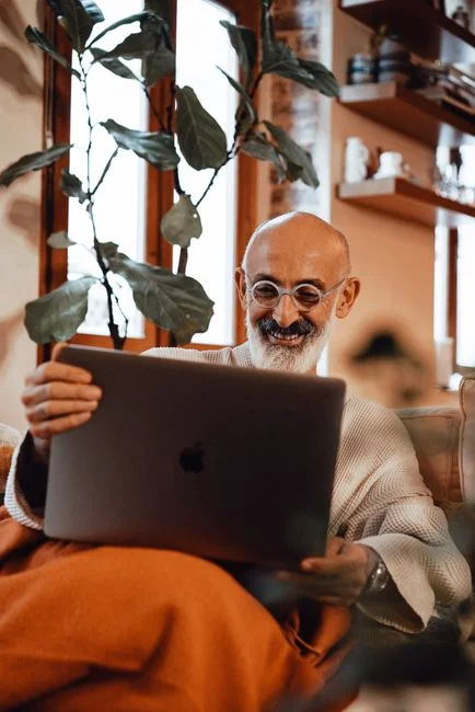 Hombre mayor sonriente con gafas teniendo una videollamada en un laptop, sentado cómodamente en interiores.