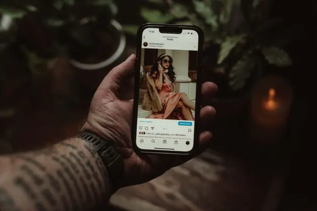 Una mano tatuada sosteniendo un smartphone que muestra una publicación de Instagram en interiores. (Foto: Pexels)
