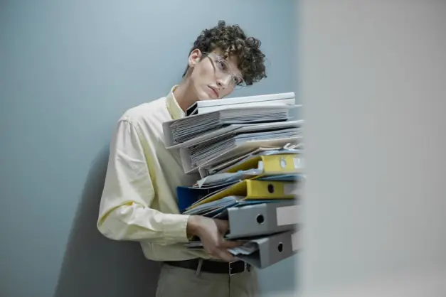 Hombre joven abrumado con una pila de carpetas simboliza el estrés laboral. (Foto: Pexels)