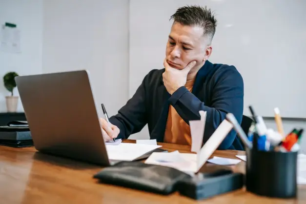 Hombre concentrado trabajando en laptop y tomando notas en un escritorio de madera en una oficina en casa. (Foto: Pexels)