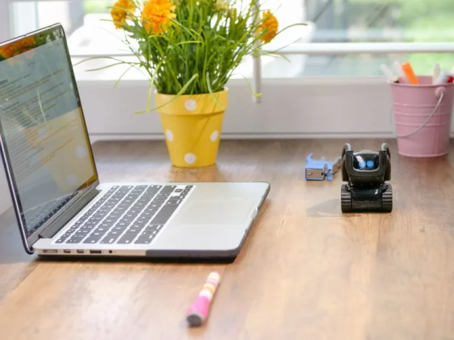 Espacio de trabajo elegante con laptop, robot de juguete y flores vibrantes con luz natural.