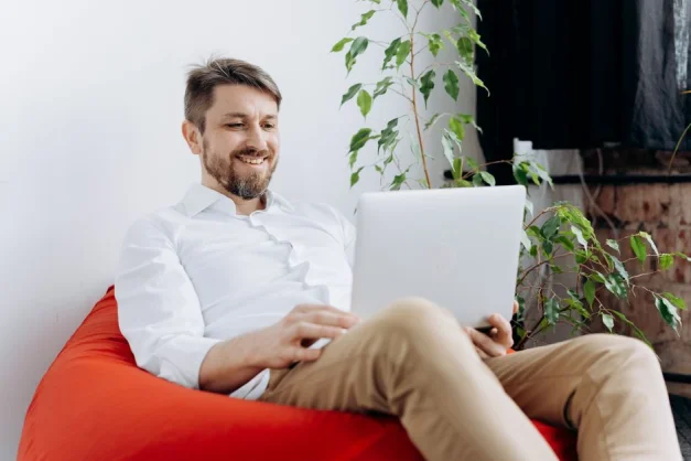Hombre sentado en una silla de frijoles roja usando un portátil, sonriendo en un espacio de trabajo moderno y casual.