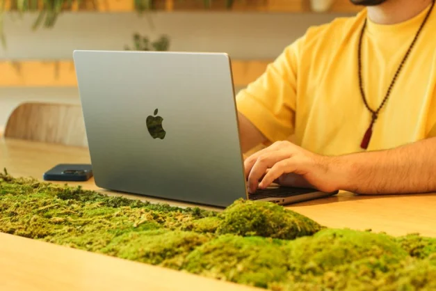 Hombre con camisa amarilla trabajando en laptop en una mesa de madera decorada con musgo en interiores.
