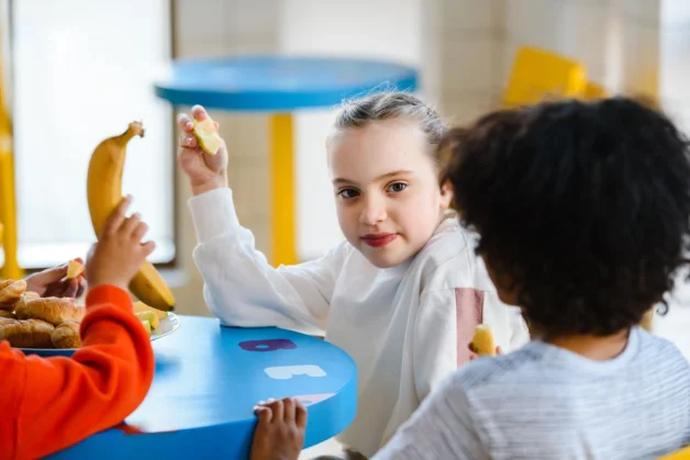 Tres niños sentados a la mesa disfrutando de plátanos y pasteles durante la merienda.