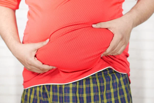 Imagen en primer plano de un hombre sosteniendo su barriga hinchada mientras lleva puesta una camisa roja.