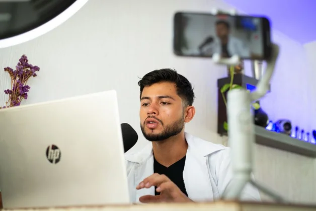 Un joven trabaja en una laptop mientras transmite contenido en línea con un teléfono inteligente.