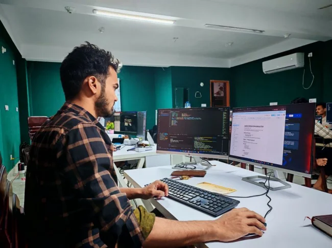 Un desarrollador de software programa en dos monitores en una oficina moderna y bien iluminada, concentrado y absorto en su trabajo.