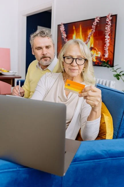 La pareja anciana sonríe mientras compra en línea con una tarjeta de crédito en un sofá acogedor.