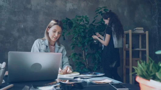 Dos mujeres trabajando en una oficina elegante con portátiles y vegetación.
