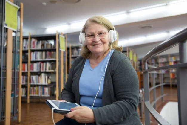 Mujer mayor sonriendo mientras escucha audiolibros en una tableta en un entorno de biblioteca.