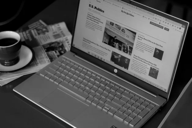 Imagen en blanco y negro de una laptop mostrando artículos de noticias, acompañada de una taza de café y periódicos.