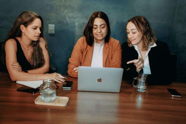 Tres mujeres trabajando juntas en un portátil, discutiendo un proyecto en un entorno profesional.