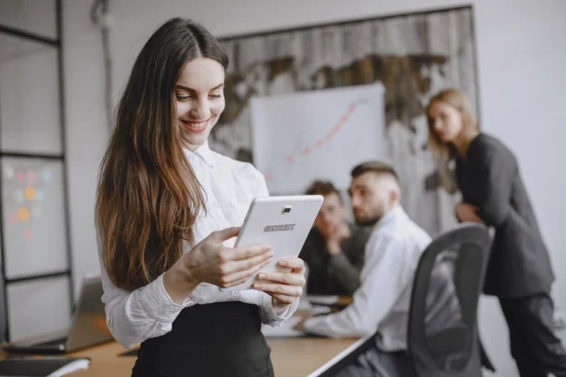 Joven mujer en un entorno de oficina profesional navegando por una tableta con colegas al fondo.