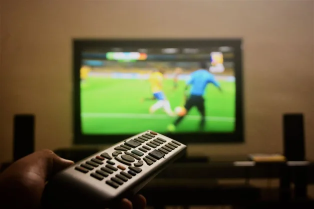 Primer plano de una mano sosteniendo un control remoto frente a una TV que muestra un partido de fútbol.