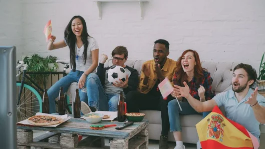 Grupo de amigos disfrutando de un partido de fútbol con bocadillos, banderas y vítores.