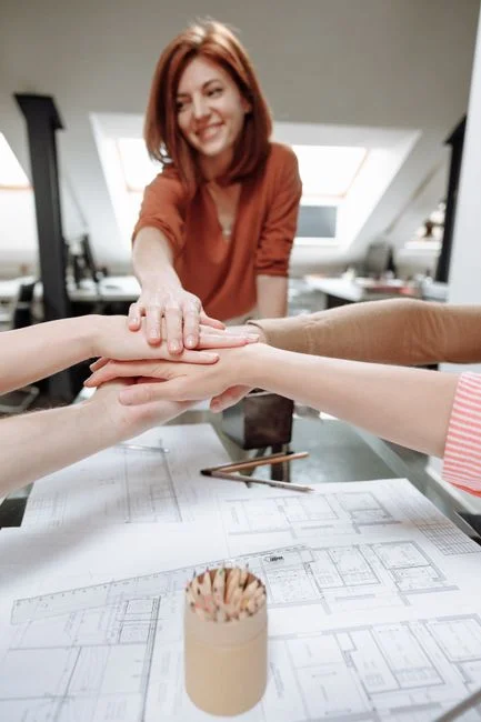 Un grupo de profesionales uniendo las manos sobre planos de escritorio en una oficina moderna, simbolizando el trabajo en equipo.