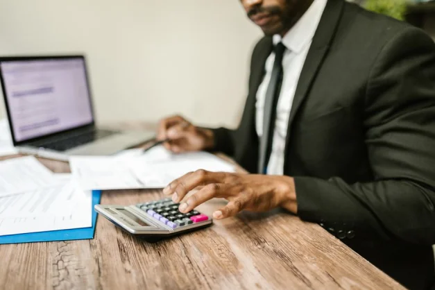 Un empresario con traje negro usando una calculadora en un escritorio con documentos financieros y un portátil.