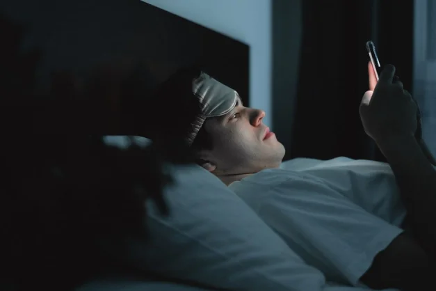 Un hombre con una máscara para dormir usando un smartphone en su cama por la noche.