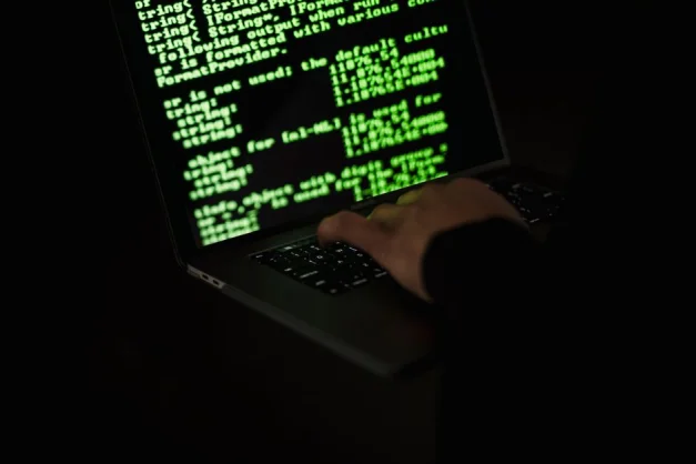 Silueta de un hacker escribiendo en un laptop en la oscuridad con código verde en la pantalla.
