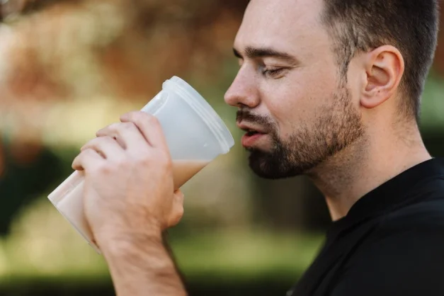 Hombre con barba bebiendo un batido de proteínas al aire libre en un día soleado, mostrando un estilo de vida saludable.